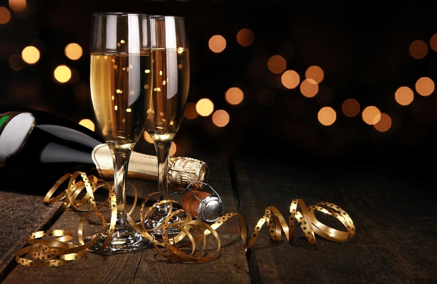 Zwei gefüllte Sektgläser neben einer geöffneten Sektflasche und goldenen Luftschlangen auf dunklem Holztisch vor unscharfen Lichtern